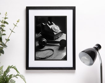 May include: Black and white photograph of a vintage rotary dial telephone with a black receiver and cord. The phone is on a wooden surface with a beam of light shining on it. The photo is framed in a black frame with a white mat.