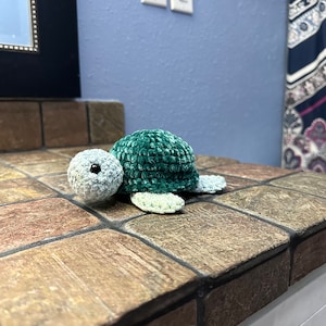 May include: A handmade crochet turtle in shades of green and gray. The turtle has a textured shell, a small head with black eyes, and four flipper-like appendages. The plush toy is displayed on a brown tiled surface.