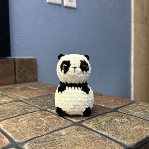 May include: A small crocheted panda bear toy with black and white fur. The panda is sitting on a tiled surface.