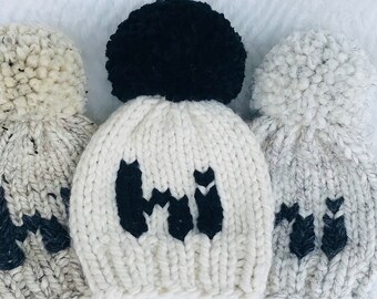 May include: Three knitted hats with pom-poms. The hats are white with black "Hi" lettering. The pom-poms are black, white with black flecks, and white.