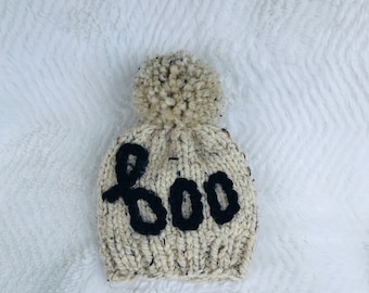 May include: A cream-colored knitted beanie with a black pom-pom and the word "BOO" knitted in black on the front.