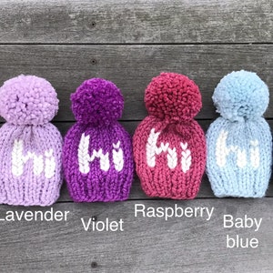 May include: Six knitted baby hats in different colors with a white pom-pom on top. The hats are pink, lavender, violet, raspberry, baby blue, and navy blue. Each hat has the word "Hi" knitted on the front in white.