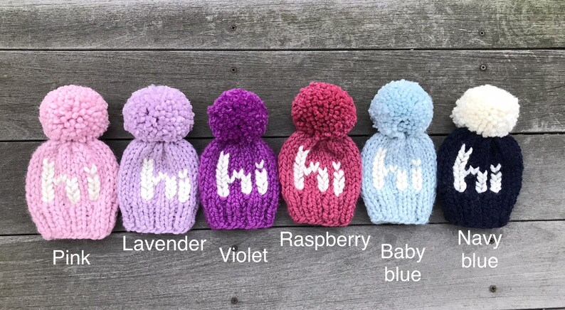 May include: Six knitted baby hats in different colors with a white pom-pom on top. The hats are pink, lavender, violet, raspberry, baby blue, and navy blue. Each hat has the word "Hi" knitted on the front in white.
