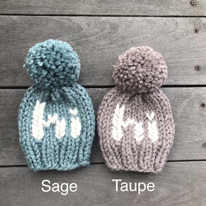 May include: Two knitted hats with pom-poms, one in sage green and one in taupe, both with white knit "Hi" lettering.
