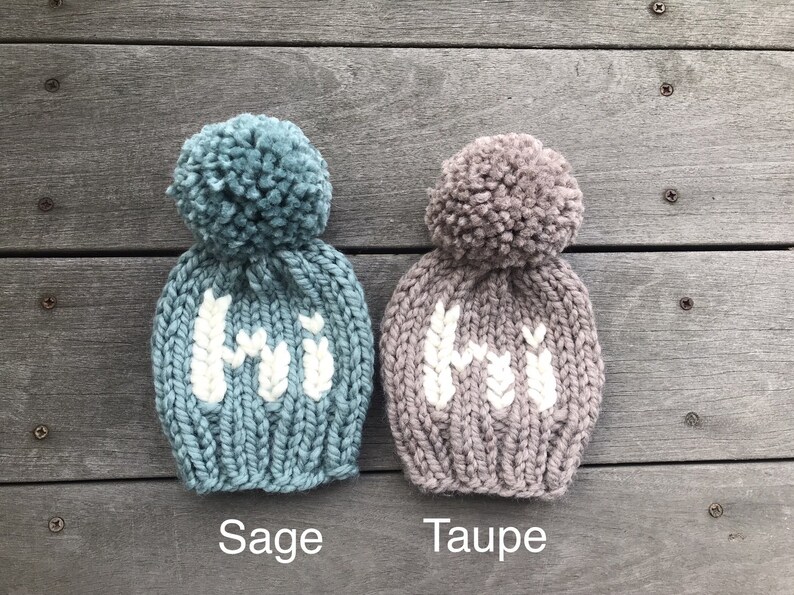 May include: Two knitted hats with pom-poms, one in sage green and one in taupe, both with white knit "Hi" lettering.