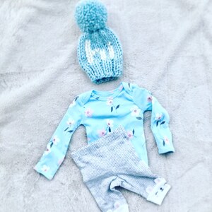 May include: A baby outfit set in blue and gray. The set includes a long-sleeved onesie with a floral print, gray striped pants with bear paw prints on the ankles, and a blue knit hat with a white pom-pom.