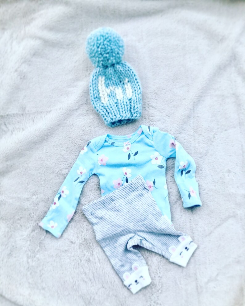 May include: A baby outfit set in blue and gray. The set includes a long-sleeved onesie with a floral print, gray striped pants with bear paw prints on the ankles, and a blue knit hat with a white pom-pom.