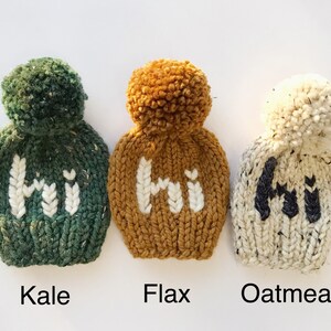 May include: Three knitted hats with pom-poms, each featuring the word "Hi" in a different color. The hats are in the colors green, mustard yellow, and white. The hats are labeled "Kale", "Flax", and "Oatmeal".