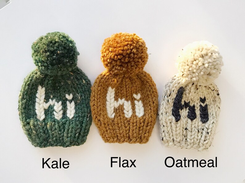 May include: Three knitted hats with pom-poms, each featuring the word "Hi" in a different color. The hats are in the colors green, mustard yellow, and white. The hats are labeled "Kale", "Flax", and "Oatmeal".