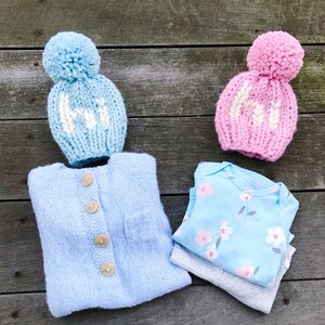 May include: Two knitted baby hats with pom-poms, one blue with white lettering and one pink with white lettering, along with a blue knitted cardigan and a blue and white floral baby onesie.