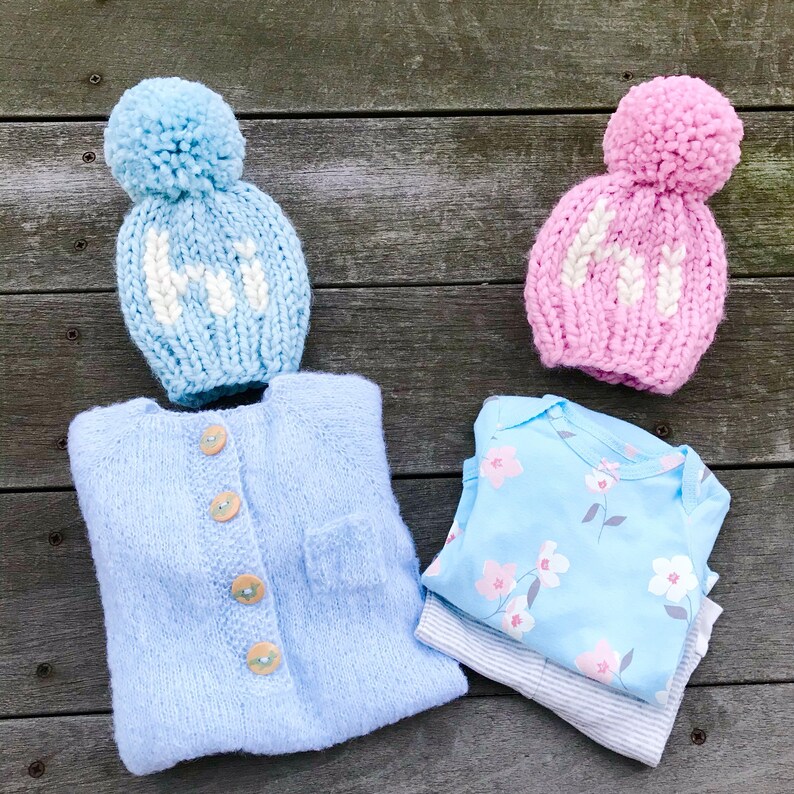 May include: Two knitted baby hats with pom-poms, one blue with white lettering and one pink with white lettering, along with a blue knitted cardigan and a blue and white floral baby onesie.