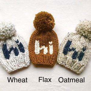 May include: Three knitted hats with pom-poms, each with the word "Hi" embroidered on the front. The hats are in the colors wheat, flax, and oatmeal.