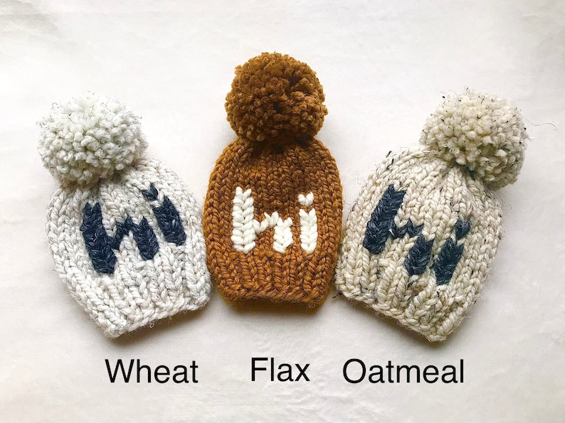 May include: Three knitted hats with pom-poms, each with the word "Hi" embroidered on the front. The hats are in the colors wheat, flax, and oatmeal.