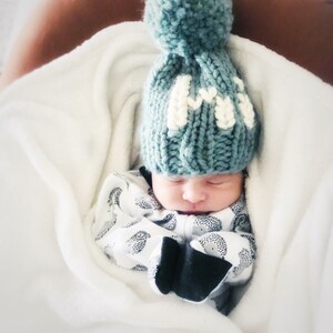 May include: A baby wearing a teal knitted hat with a white pom pom and the letter "M" embroidered on the front. The baby is sleeping in a white blanket.