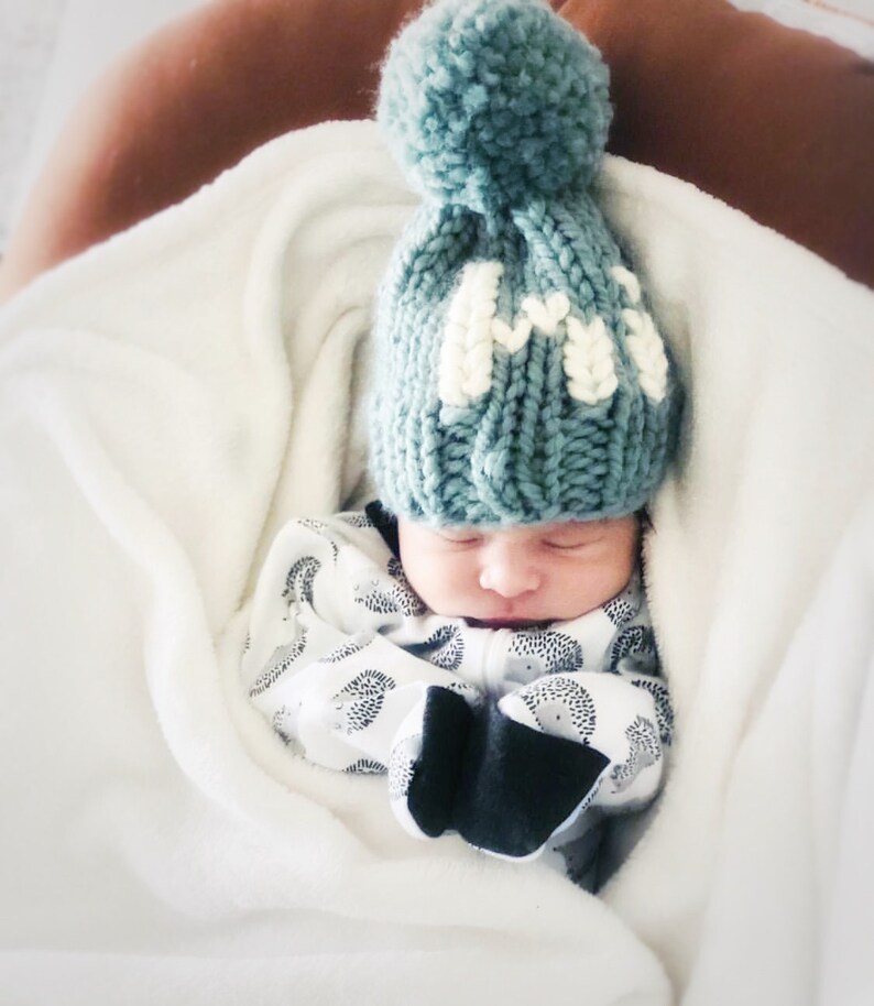 May include: A baby wearing a teal knitted hat with a white pom pom and the letter "M" embroidered on the front. The baby is sleeping in a white blanket.