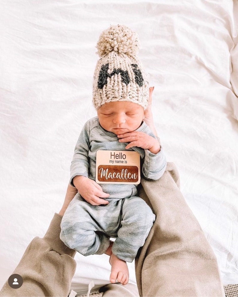 May include: A newborn baby wearing a beige knitted hat with a pom-pom and black lettering that says "HI". The baby is holding a wooden sign that says "Hello my name is Macallen".