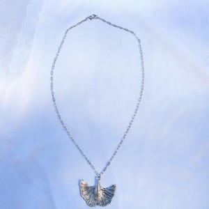 May include: A silver-toned necklace featuring a ginkgo leaf pendant. The pendant has detailed veining, and the chain is a delicate link style. The necklace is displayed on a white, textured background, creating a simple and elegant presentation.