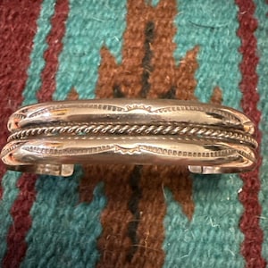 May include: A silver cuff bracelet with a twisted rope design in the center. The bracelet has etched details and is displayed on a turquoise, brown, and red patterned textile.