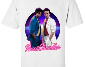 May include: White t-shirt featuring a graphic of two men in retro attire, set against a purple and blue background. The text "Ross & Chandler" is displayed in pink script below the image. The shirt is made of a soft material.