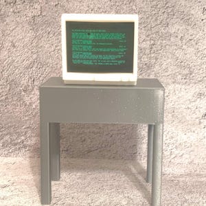 May include: A small gray table with a white computer monitor on top. The monitor displays green text on a black background.