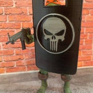 May include: A green action figure of a Teenage Mutant Ninja Turtle holding a black machine gun and a black shield with a silver skull emblem. The figure is posed in front of a brick wall.