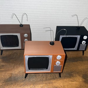 May include: Three miniature television sets in brown, orange, and black. Each unit features a silver screen frame, two control knobs, a vertical speaker grille, and a thin wire antenna extending from the top. The sets stand on four small legs.