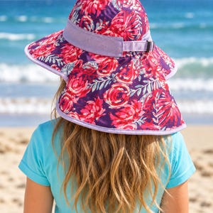 May include: A sun hat featuring a vibrant floral pattern in shades of deep purple, bright pink, and red. The hat includes a wide brim, a light purple mesh band with an adjustable strap, and a light purple trim along the edges of the brim.