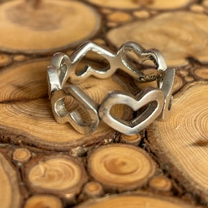 May include: A silver ring with a design of interlocking hearts. The ring is on a wooden surface.
