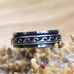 May include: A silver spinning ring with a wave design. The ring is on a textured surface.