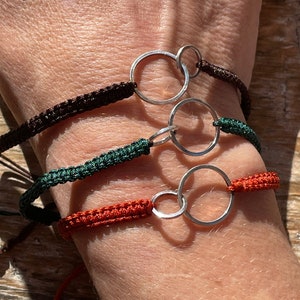 May include: Three bracelets with different colored cords and silver double circle charms. The cords are brown, green, and orange.