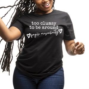 Peut inclure: Un t-shirt noir avec un texte blanc qui dit "trop maladroit pour être autour de la masculinité fragile" avec un symbole de cœur de chaque côté du texte.