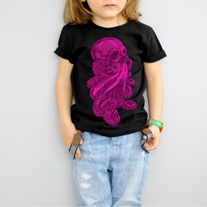 Puede incluir: Un niño o niña lleva una camiseta negra con un gráfico de un pulpo rosa. El niño o niña lleva pantalones vaqueros azules y una pulsera verde y naranja.