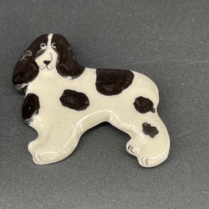May include: A ceramic figurine of a black and white spotted dog. The dog is standing on all four legs and has its head tilted to the side. The figurine is painted in a realistic style.