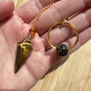 Op de afbeelding: Een goudkleurige pendel met een tijgeroogsteen en een gouden ketting. De pendel wordt in een hand gehouden. Een gouden ring met een zwart-bruin gespikkelde steen is ook zichtbaar.