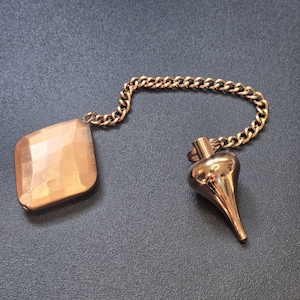 May include: A copper-toned metal plumb bob with a smooth, pointed tip connected by a matching link chain to a faceted, diamond-shaped copper-toned weight. The components feature a polished, reflective finish and are linked together by a small metal ring.