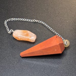 Puede incluir: Un péndulo con un colgante de piedra roja facetada en forma de cono unido a una cadena de metal plateado. Una cuenta de piedra lisa, translúcida y de color naranja claro también está unida a la cadena, junto con una pequeña cuenta amarilla.
