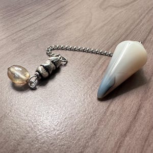 Puede incluir: Un péndulo con un cristal blanco en forma de lágrima y una cadena plateada. La cadena se conecta a una pieza decorativa plateada y una pequeña cuenta de color ámbar. El cristal tiene un sutil tono azul en la punta.