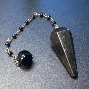 May include: A black faceted stone pendulum with a pointed tip attached to a gold-toned metal chain. The chain features small black beads and ends with a single smooth, rounded black stone bead, creating a decorative and functional hanging object.