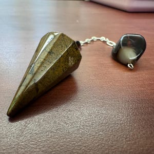 May include: A faceted, cone-shaped pendulum made of brown and tan stone, connected by a silver chain to a rounded, dark stone. The pendulum is designed for divination and spiritual practices.