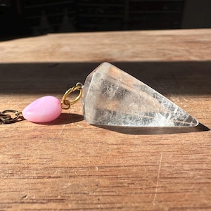 Könnte beinhalten: Ein klares Quarz-Kristallpendel mit einer rosa tropfenförmigen Perle und einer goldfarbenen Kette. Der Kristall ist facettiert und verjüngt sich zu einer Spitze. Das Pendel wird für die Wahrsagung und spirituelle Praktiken verwendet.
