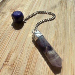 May include: A purple and white crystal pendulum with a silver chain and a dark purple sphere. The crystal is a hexagonal shape. The pendulum is on a wooden surface.
