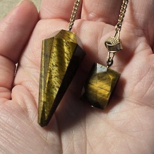 Puede incluir: Un péndulo de piedra ojo de tigre facetada y puntiaguda junto a un dije de ojo de tigre rectangular facetado, ambos suspendidos de una cadena de metal dorado. Las piedras presentan bandas chatoyantes de color marrón dorado.