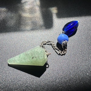 Puede incluir: Un péndulo de piedra verde claro facetado unido a una cadena de metal plateado. La cadena presenta dos cuentas de vidrio decorativas, una en azul real profundo y otra en azul cielo más claro, conectadas a la parte superior del colgante.