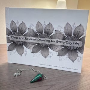 Può includere: Un libro intitolato "Clear and Release: Dowsing for Every Day Life" con illustrazioni floreali e un pendolo. Il pendolo ha una pietra preziosa verde e rossa e una catena d'argento. Il libro è una guida alla rabdomanzia per principianti.