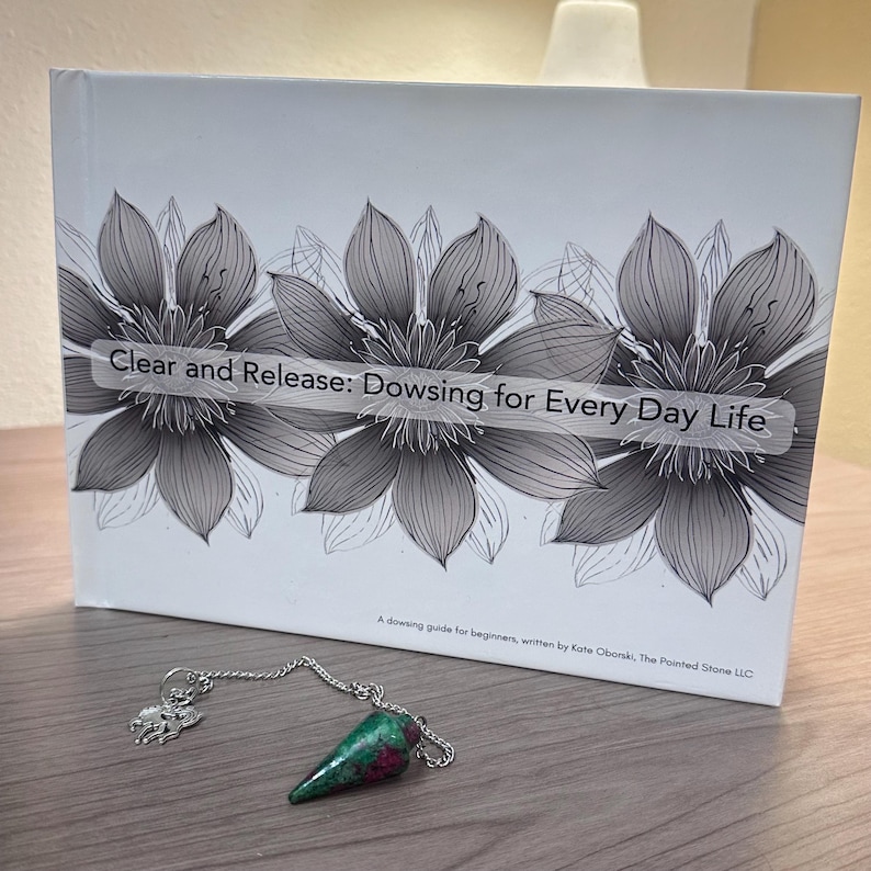 May include: A book titled "Clear and Release: Dowsing for Every Day Life" with floral illustrations and a pendulum. The pendulum has a green and red gemstone and a silver chain. The book is a dowsing guide for beginners.