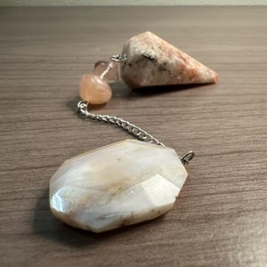 May include: A pendulum with a faceted, light-colored stone at the bottom, connected by a silver chain to a few smaller, rounded stones and a larger, triangular stone. The stones have a mix of peach and white tones.