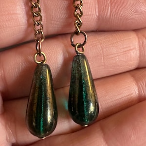 Puede incluir: Dos colgantes de cristal en forma de lágrima con un acabado iridiscente en verde oscuro y bronce, cada uno suspendido de un eslabón de cadena metálico en tono bronce. Los colgantes presentan una superficie lisa y reflectante con variaciones.