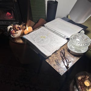 Puede incluir: Una vela encendida junto a una carpeta abierta con notas manuscritas y un par de gafas en una mesita. Un plato decorativo de cristal con tapa descansa sobre un pequeño soporte blanco. También se ve un jarrón con flores secas.