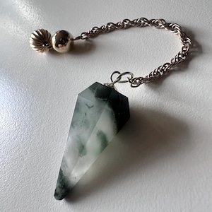 May include: A faceted crystal pendulum with a silver chain and accents. The crystal is a mix of clear and green hues. The chain connects to a silver bead and a decorative, ridged silver piece.