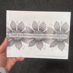 May include: A white book with the title "Clear and Release: Dowsing for Every Day Life" is held in a hand. The cover features three large, grey floral illustrations. Below the title is the text "A dowsing guide for beginners, written by Kate Oborski, The Pointed Stone LLC."